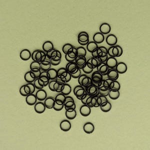 May include: A collection of small, black metal jump rings scattered on a light green surface. The rings are circular and appear to be made of a durable material, suitable for jewelry making or crafts. The image is a top-down view.
