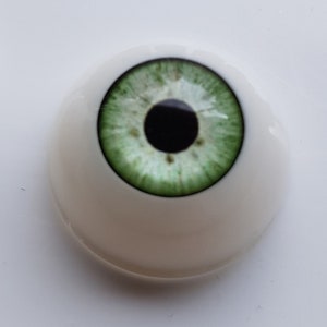Puede incluir: Un solo ojo prot&eacute;sico de aspecto realista con un iris verde claro y una pupila negra. El ojo est&aacute; colocado en una base blanca y redondeada, dise&ntilde;ada para imitar la apariencia natural de un ojo humano. El iris tiene vetas detalladas.