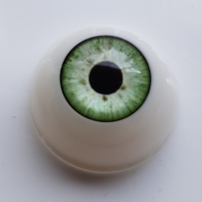Puede incluir: Un solo ojo prot&eacute;sico de aspecto realista con un iris verde claro y una pupila negra. El ojo est&aacute; colocado en una base blanca y redondeada, dise&ntilde;ada para imitar la apariencia natural de un ojo humano. El iris tiene vetas detalladas.