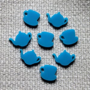 Könnte beinhalten: Eine Sammlung von sieben flachen, soliden blauen Acryl-Anhängern in Form von Teekannen und Teetassen. Es gibt drei teekannenförmige und vier tassenförmige Anhänger, die alle eine glatte, opake Oberfläche und eine einheitliche leuchtend blaue Farbe aufweisen.