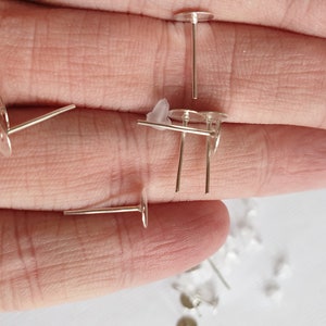 May include: Silver earring posts with flat round backs. The posts are laying on a hand.