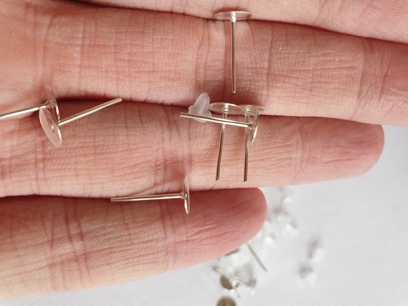 May include: Silver earring posts with flat round backs. The posts are laying on a hand.