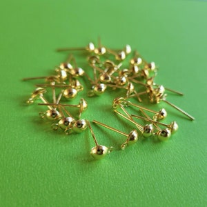 May include: A pile of gold-coloured earring posts with small round heads. The posts are scattered on a green surface.