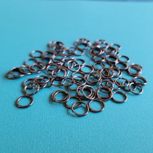 Könnte beinhalten: Eine Ansammlung kleiner, dunkelgrauer Metall-Binderinge auf einer leuchtend türkisfarbenen Oberfläche. Die Ringe sind kreisförmig und scheinen aus einem haltbaren Metall gefertigt zu sein, das sich für die Schmuckherstellung oder Bastelarbeiten eignet. Das Bild ist gut beleuchtet.