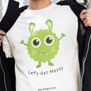 May include: White t-shirt with a green cartoon monster graphic and the text "Let's Get Messy" and "The Single Life". The shirt is being worn with a black jacket and sunglasses.