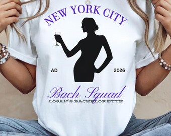 May include: White t-shirt with a purple design. The design includes the text "NEW YORK CITY" above a black silhouette of a person holding a wine glass. Below the silhouette is the text "Bach Squad" and "LOGAN'S BACHELORETTE".