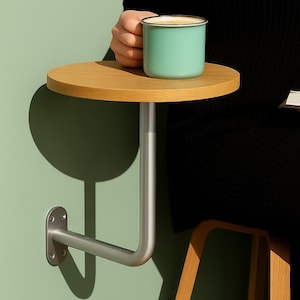 Pode incluir: Uma mesa lateral montada na parede com um tampo redondo de madeira clara. A mesa é suportada por um braço e um suporte de metal prateado. Uma caneca turquesa com uma borda prateada está sobre a mesa. A mesa está fixada a uma parede verde claro.