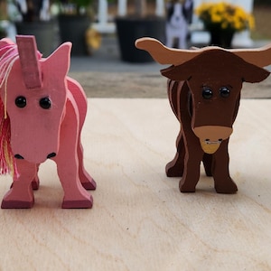 May include: Wooden animal figurines including a white unicorn with a gold horn, a pink unicorn, and two brown longhorn cattle. The figurines are on a light wood surface.