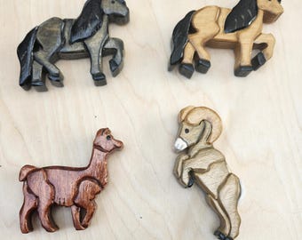 Könnte beinhalten: Vier Holztiere: zwei Pferde, ein Lama und ein Widder. Die Pferde sind in Grau- und Beigetönen mit schwarzen Mähnen bemalt. Das Lama ist braun und der Widder ist beige mit weißen Akzenten. Alle befinden sich auf einer hellen Holzoberfläche.