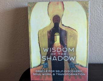 May include: A rectangular oracle card deck box featuring a dark silhouette of a figure standing in a yellow archway. The text reads WISDOM OF THE SHADOW and ORACLE FOR SELF-DISCOVERY, SOUL WORK, & TRANSFORMATION. The figure has blue-toned hands.