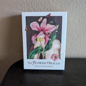 May include: A rectangular box for The Flower Oracle 44 Card Deck & Guidebook. The front features a central illustration of a large pink orchid with green leaves, surrounded by small butterflies, set against a dark background with a human figure.