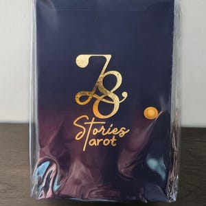 May include: A tarot deck in a clear plastic wrap. The deck has a navy blue background with a gold foil design. The design includes the text "7 Stories tarot". The deck is rectangular.