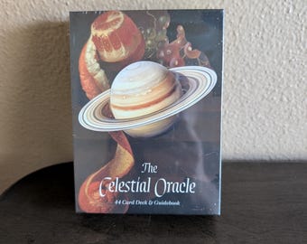 May include: A rectangular box for The Celestial Oracle 44 Card Deck & Guidebook. The front features a graphic of the planet Saturn with rings, a peeled orange, and grapes against a dark background. The text reads The Celestial Oracle 44 Card Deck & Guidebook.