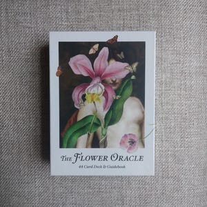 May include: A white box featuring "The Flower Oracle" with a pink orchid illustration and butterflies. The text "44 Card Deck & Guidebook" is below the title. The image has a dark background with a person's face partially visible.