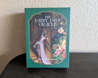 May include: A rectangular green box for The Fairy Tale Oracle 44 Card Deck & Guidebook. The front features an illustration of a figure in a long pink dress and pointed hat standing on stairs, next to a vibrant floral arrangement of roses and greenery.