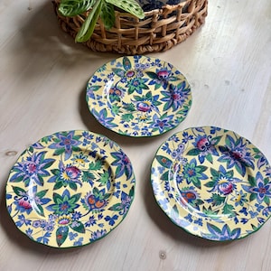 May include: Three round ceramic plates with scalloped edges, featuring a vibrant yellow background decorated with a dense, colorful floral pattern in shades of blue, green, purple, and orange. The plates are arranged on a light wooden surface.