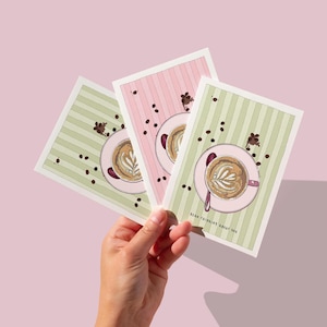 May include: Three greeting cards with coffee art are held against a pink background. Each card features a latte design on a pink saucer, surrounded by coffee beans. The cards have striped backgrounds in pink and green. The text "BEAN THINKING ABOUT YOU" is visible.
