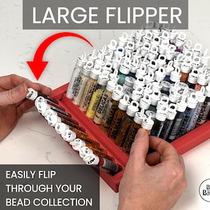 May include: A red plastic storage tray containing multiple clear cylindrical tubes filled with colorful beads, each topped with a white cap. The tray features a hinged section labeled LARGE FLIPPER, allowing users to easily flip through the bead collection.