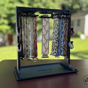 Könnte beinhalten: Ein schwarzer Präsentationsständer mit mehreren Armbändern mit verschiedenen Mustern und Farben. Die Armbänder hängen von der Oberseite des Ständers, wobei Designs mit roten, weißen und goldenen Akzenten gezeigt werden. Der Ständer steht auf einer dunklen Oberfläche.