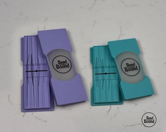 May include: Two rectangular needle cases, one lavender and one teal, each featuring a sliding cover with a circular logo reading Bead Bound. The cases contain thin metal needles held in place by a central horizontal bar with a small black dot.