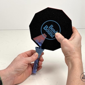 Könnte beinhalten: Eine Person schüttet blaue Perlen in eine schwarze achteckige Schale mit dem aufgedruckten Logo "Bead Bound".