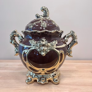 May include: A brown ceramic serving dish with gold accents and ornate details. The dish has a lid and two handles. It is decorated with floral and scroll designs.