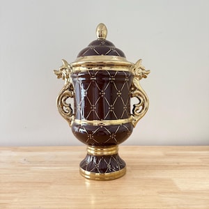 May include: A decorative, lidded urn with a dark brown base and gold accents. The urn features a diamond pattern with gold detailing, ornate gold handles, and a gold finial on the lid. The base and rim are also trimmed in gold.