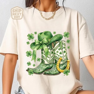 Puede incluir: Camiseta color crema con un diseño de acuarela de un sombrero de vaquero verde con un lazo, botas de vaquero verdes, una herradura dorada y tréboles verdes. La camiseta tiene el texto "SHIP NEXT DAY" en la esquina superior izquierda.