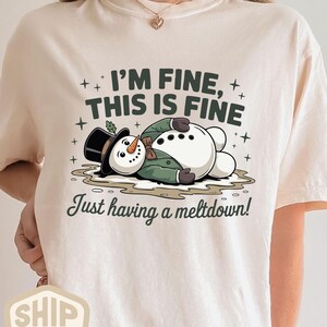 Może przedstawiać: Beżowa koszulka z kreskówkowym bałwanem topniejącym na ziemi. Bałwanek ma czarny cylinder i zieloną kurtkę. Tekst na koszulce brzmi "I'm fine, this is fine" i "Just having a meltdown!"