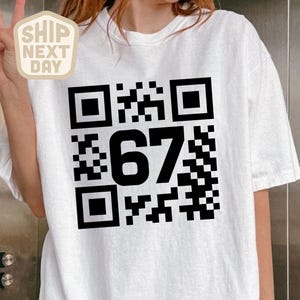 Puede incluir: Camiseta blanca con un gran código QR negro y el número "67" impreso en la parte delantera. La camiseta tiene mangas cortas y un corte holgado. Una pegatina de "Envío al día siguiente" es visible.
