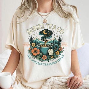 May include: Cream-colored t-shirt featuring a teal teacup graphic with steam rising to a crescent moon and stars. The design includes text that reads "SURIEL TEA CO" and "The Hottest Tea in Prythian."