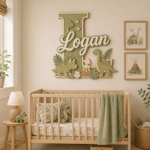 May include: A large wooden wall sign featuring the letter L in sage green with the name Logan in white script. The sign is decorated with cutouts of dinosaurs, a volcano, and an egg. Two framed dinosaur prints hang on the wall nearby.