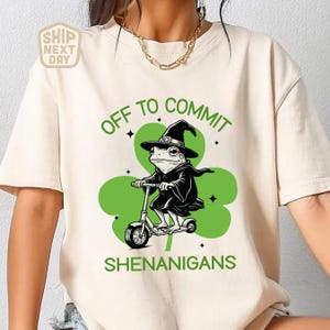 May include: Beige t-shirt with a green shamrock design and the text "OFF TO COMMIT SHENANIGANS." The graphic features a frog in a witch hat riding a scooter. The shirt has a round neckline and short sleeves.