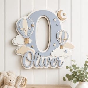 Pode incluir: Uma placa de parede de madeira em camadas com uma letra O grande em azul claro, o nome Oliver em letra cursiva, dois balões de ar quente, nuvens, estrelas e uma lua crescente em tons de azul, bege e branco com acabamento fosco.