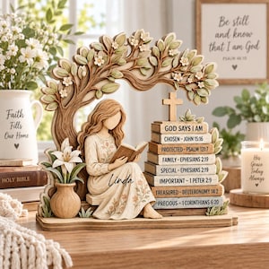 May include: A decorative figurine featuring a person reading a book under a tree with white flowers. Beside them is a stack of books labeled with religious text including 'GOD SAYS I AM' and 'CHOSEN', topped with a wooden cross and a small vase.