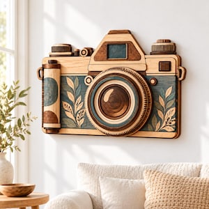 May include: A wall-mounted wooden camera sculpture featuring layered light and dark wood tones. The design includes a central circular lens, a film roll on the left, and decorative leaf patterns in blue and cream hues on the front panel.