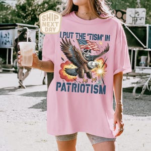 Puede incluir: Camiseta rosa claro con el texto "I PUT THE 'TISM IN PATRIOTISM" y un gr&aacute;fico de un &aacute;guila que lleva un mapache con una bandera estadounidense. El &aacute;guila est&aacute; rodeada de explosiones rojas y amarillas.