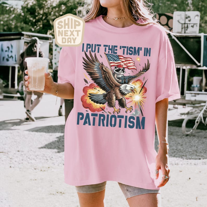 Puede incluir: Camiseta rosa claro con el texto "I PUT THE 'TISM IN PATRIOTISM" y un gr&aacute;fico de un &aacute;guila que lleva un mapache con una bandera estadounidense. El &aacute;guila est&aacute; rodeada de explosiones rojas y amarillas.