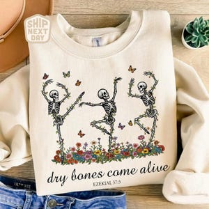 Puede incluir: Sudadera color crema con un gráfico de tres esqueletos bailando con detalles florales y mariposas. La frase "dry bones come alive" está impresa debajo de los esqueletos, con el versículo bíblico Ezequiel 37:5. Un diseño divertido y único.