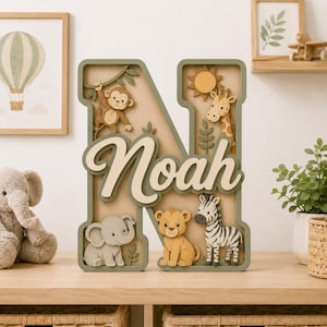 以下が含まれることがあります： 筆記体でNoahという名前が書かれた、層状の大きな木製の文字N。デザインには、サル、キリン、ゾウ、ライオン、シマウマの切り抜きフィギュアに加え、太陽と葉が含まれており、すべてベージュ、茶色、緑の色合いで構成されています。