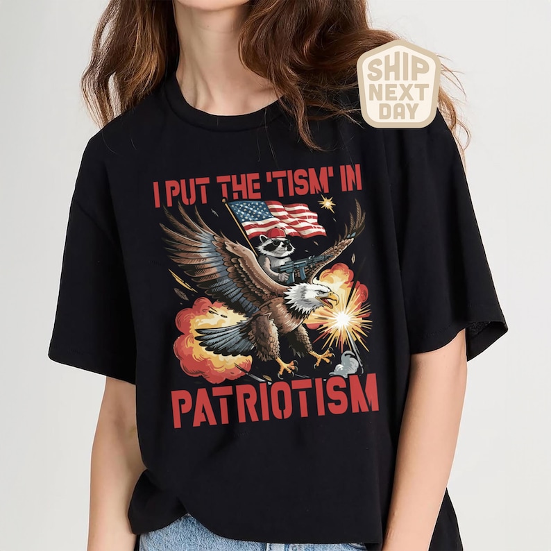 Puede incluir: Camiseta negra con un gr&aacute;fico que presenta un &aacute;guila que lleva un mapache con una bandera estadounidense y una ametralladora. El texto dice "I PUT THE 'TISM' IN PATRIOTISM" en rojo.
