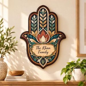 Peut inclure: Une plaque murale en forme de main de Hamsa avec un cadre en bois foncé et des découpes superposées complexes. Le motif présente des éléments floraux bleu sarcelle, orange et crème avec un panneau central en bois clair portant le texte The Khan Family.