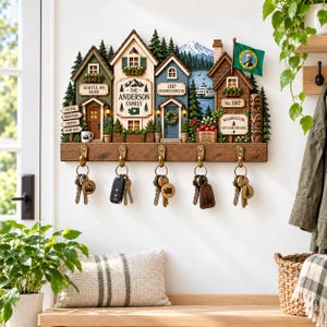 May include: A wall-mounted wooden key holder featuring a miniature village scene with houses, evergreen trees, and a mountain backdrop. It includes five brass hooks holding keys, personalised signs with the text The Anderson Family, and a Washington state flag.