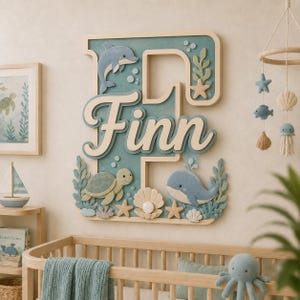 Könnte beinhalten: Ein großes Wandschild aus Holz mit dem Buchstaben F in gedecktem Blaugrün, überlagert mit dem Namen Finn in weißer Schreibschrift. Das Schild ist mit Ausschnitten eines Delfins, einer Meeresschildkröte, eines Wals und Muscheln verziert.