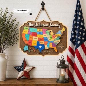 May include: A wooden wall sign featuring a colorful map of the United States with individual states labeled. The sign displays the text The Johnson Family at the top and EST. 2026 at the bottom, suspended by a rope from a black metal hook.