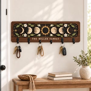 May include: A rectangular wooden wall-mounted key holder featuring a dark panel with a moon phase cycle, white flowers, and green leaves. The text THE MILLER FAMILY is printed in white. It includes six black metal hooks with keys attached.