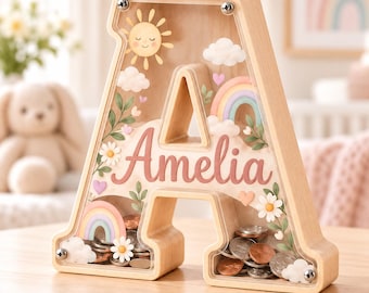 Puede incluir: Una hucha de madera con forma de letra A y panel frontal de acrílico transparente. El nombre Amelia está impreso en letra cursiva rosa. El diseño incluye un sol, nubes, arcoíris, flores y corazones. Tornillos metálicos aseguran el frente.