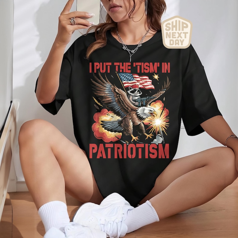 Puede incluir: Camiseta negra con la frase "I PUT THE 'TISM' IN PATRIOTISM". El dise&ntilde;o presenta un &aacute;guila llevando un mapache con una bandera y destellos de luz.