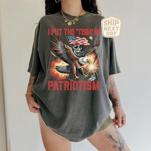 Puede incluir: Camiseta gris oscuro con un gr&aacute;fico patri&oacute;tico. El dise&ntilde;o presenta un &aacute;guila que lleva un mapache sosteniendo una bandera, con el texto "I PUT THE 'TISM' IN PATRIOTISM" en rojo. La camiseta tiene un aspecto vintage.