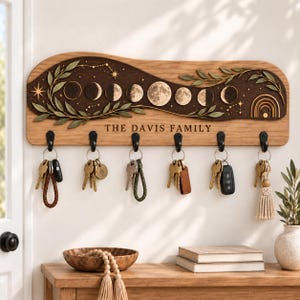 May include: A wooden wall-mounted key holder with six black metal hooks. The board features a dark wood inlay with a moon phase cycle, star motifs, and leafy branches. The text "THE DAVIS FAMILY" is engraved in the center of the light wood panel.
